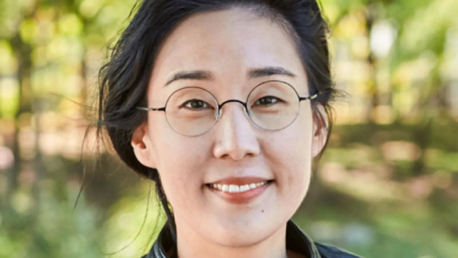Color Image of Angie Heo, an individual with light skin, dark hair, and glasses. Green trees and grass are visible in the background