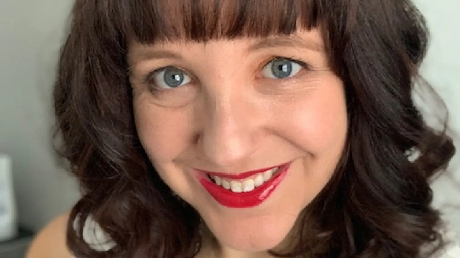 a headshot of a woman with medium length brown hair, blue eyes, and bright red lipstick smiling