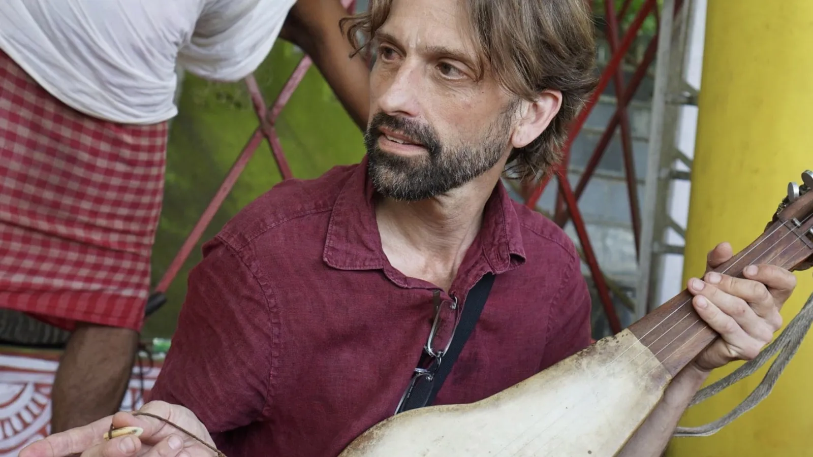 a man with short hair and a beard holds a string instrument and points to something out of the frame