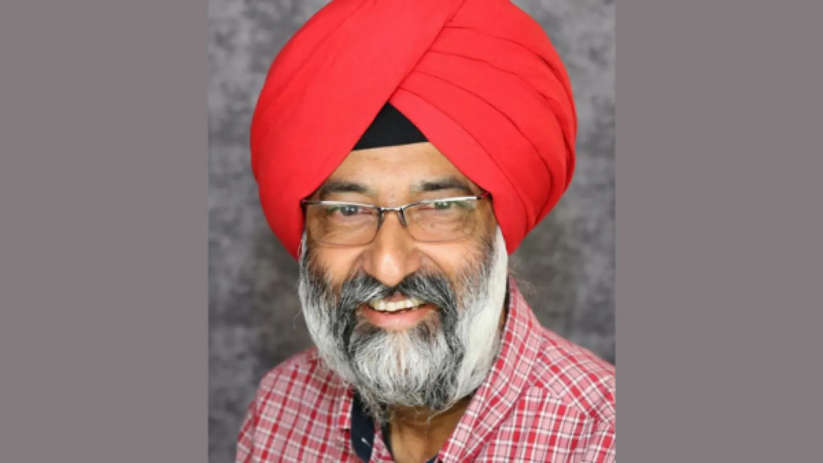 Front facing headshot of a man with a grey beard and glasses, wearing a plaid shirt and a turban on his head. He is smiling. 
