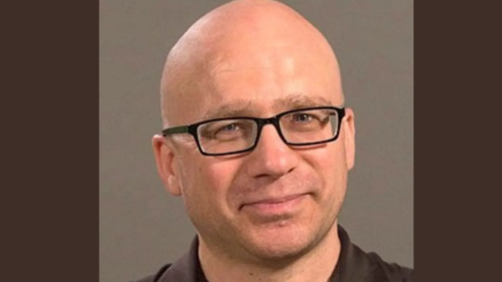 Portrait of a bald white man wearing square black glasses and a slight smile on his face. 