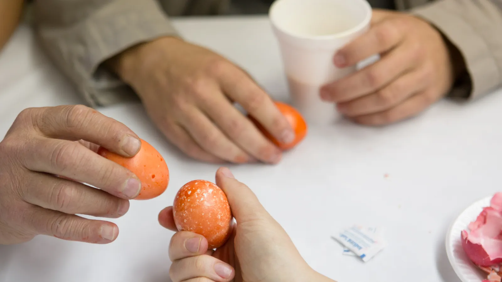 Four hands are in the image. Three hands hold an orange egg. Two eggs are nearly touching, about to crack. 