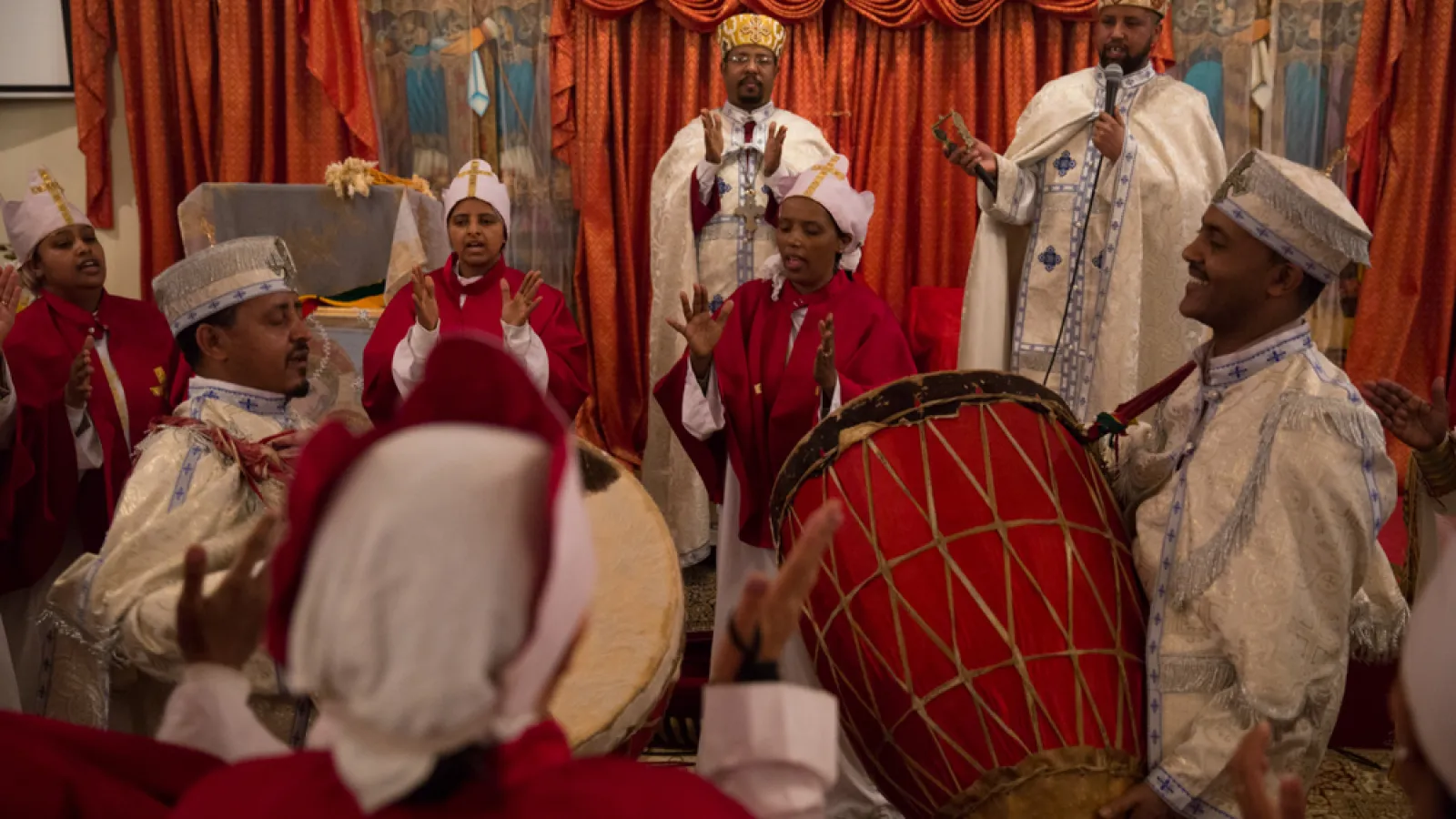 People in white gowns are playing large animal-skin drums