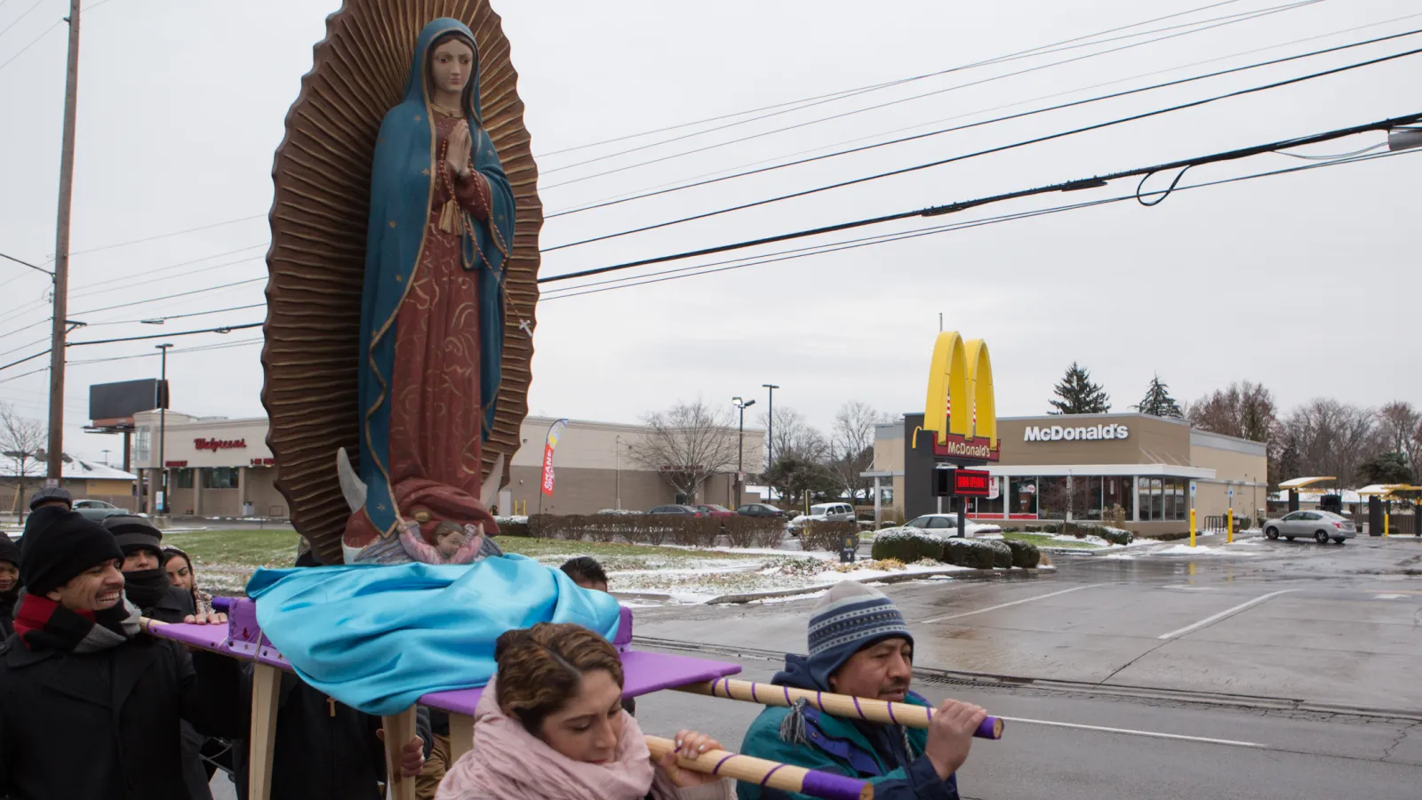 Four people hold poles that are attached to a platform. A large sculpture of Our Lady of Guadaloupe is on the platform. They are in a parking lot. 