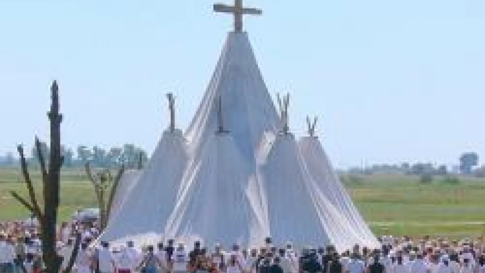 A cloth tent with five pointy steeples with a group of over one hundred people cirlcled around it. 