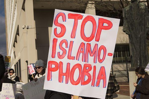 Color image of a white protest poster with red letters reading "Stop Islamophobia." A beige stone building and four individuals dressed in winter clothing are in the background.