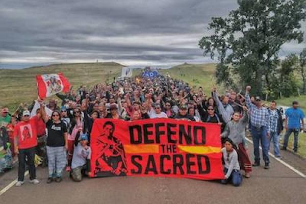 Color image of a very large group of individuals gathered to protest the DAPL behind a large red sign with a yellow stripe and black text which reads "Defend the Sacred." A vast, cloudy, grey sky is visible in the top half of the image. 