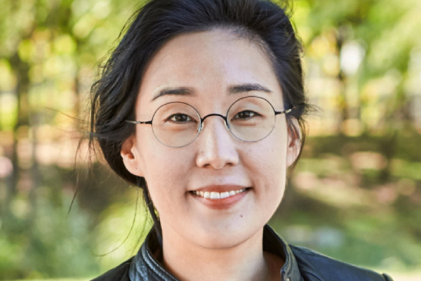 Color Image of Angie Heo, an individual with light skin, dark hair, and glasses. Green trees and grass are visible in the background