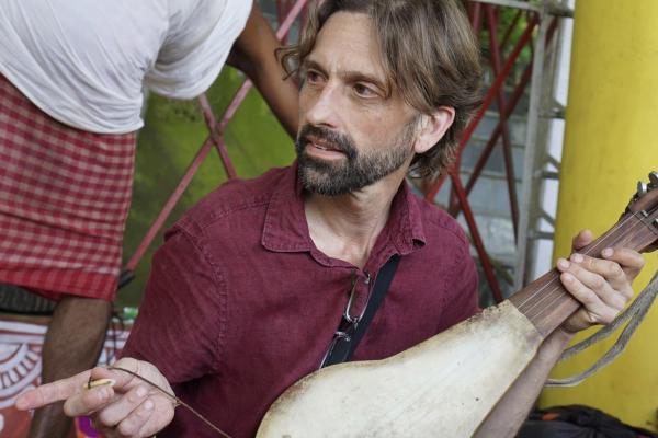 a man with short hair and a beard holds a string instrument and points to something out of the frame