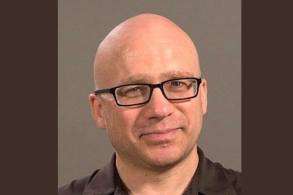 Portrait of a bald white man wearing square black glasses and a slight smile on his face. 