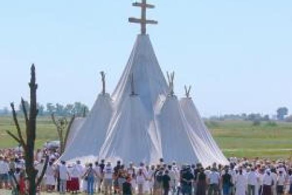 A cloth tent with five pointy steeples with a group of over one hundred people cirlcled around it. 