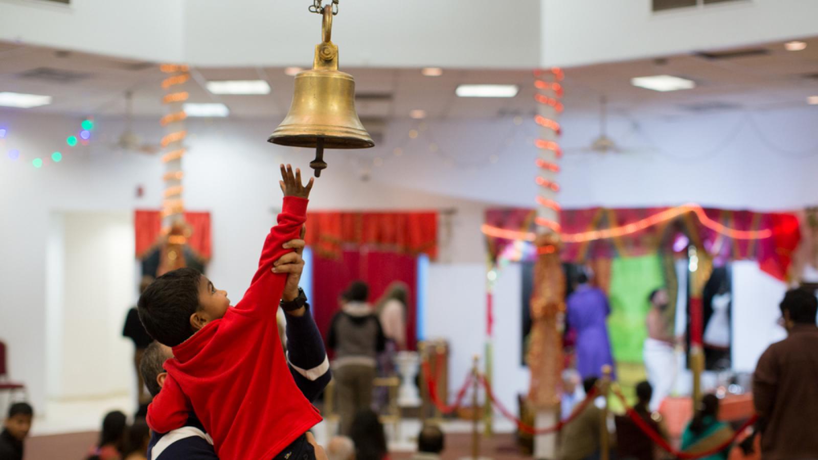 A man holds up a small boy, he is reaching to ring a large bell suspended from the ceiling