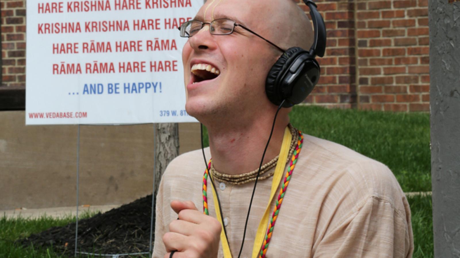 A man with a shaved head and glasses is holding a brass percussion instrument and his mouth is open like he is singing