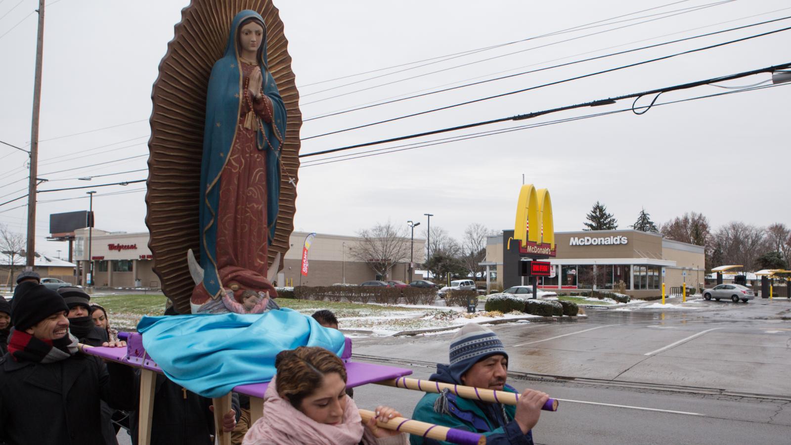 Four people hold poles that are attached to a platform. A large sculpture of Our Lady of Guadaloupe is on the platform. They are in a parking lot. 