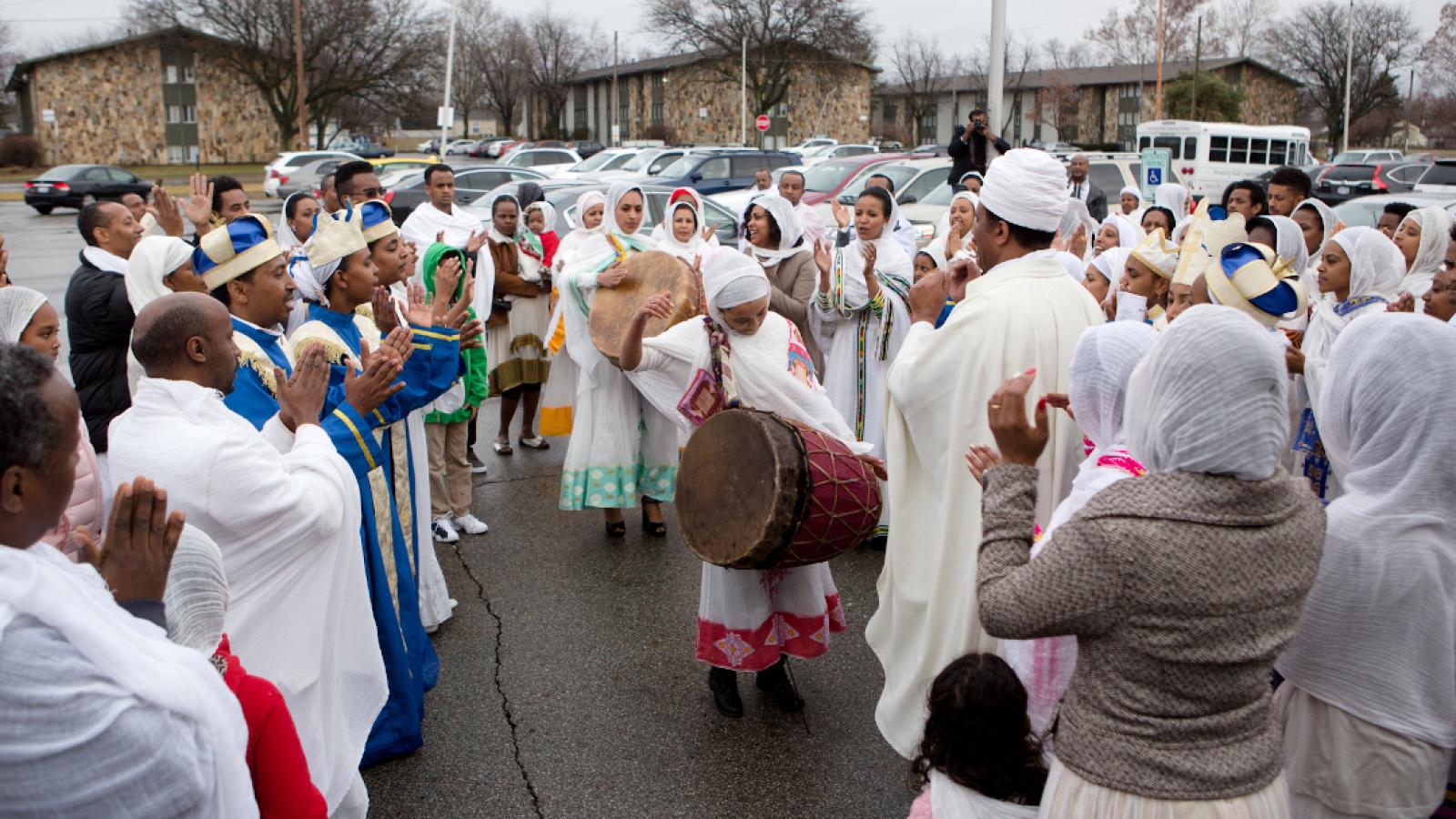 A girl plays an animal skin drum in a parking lot, surrounded by dozens of people wearing white robes